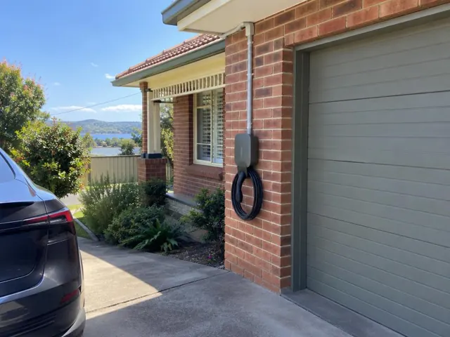 Wall-mounted EV charger on a brick garage wall beside a driveway in Teralba